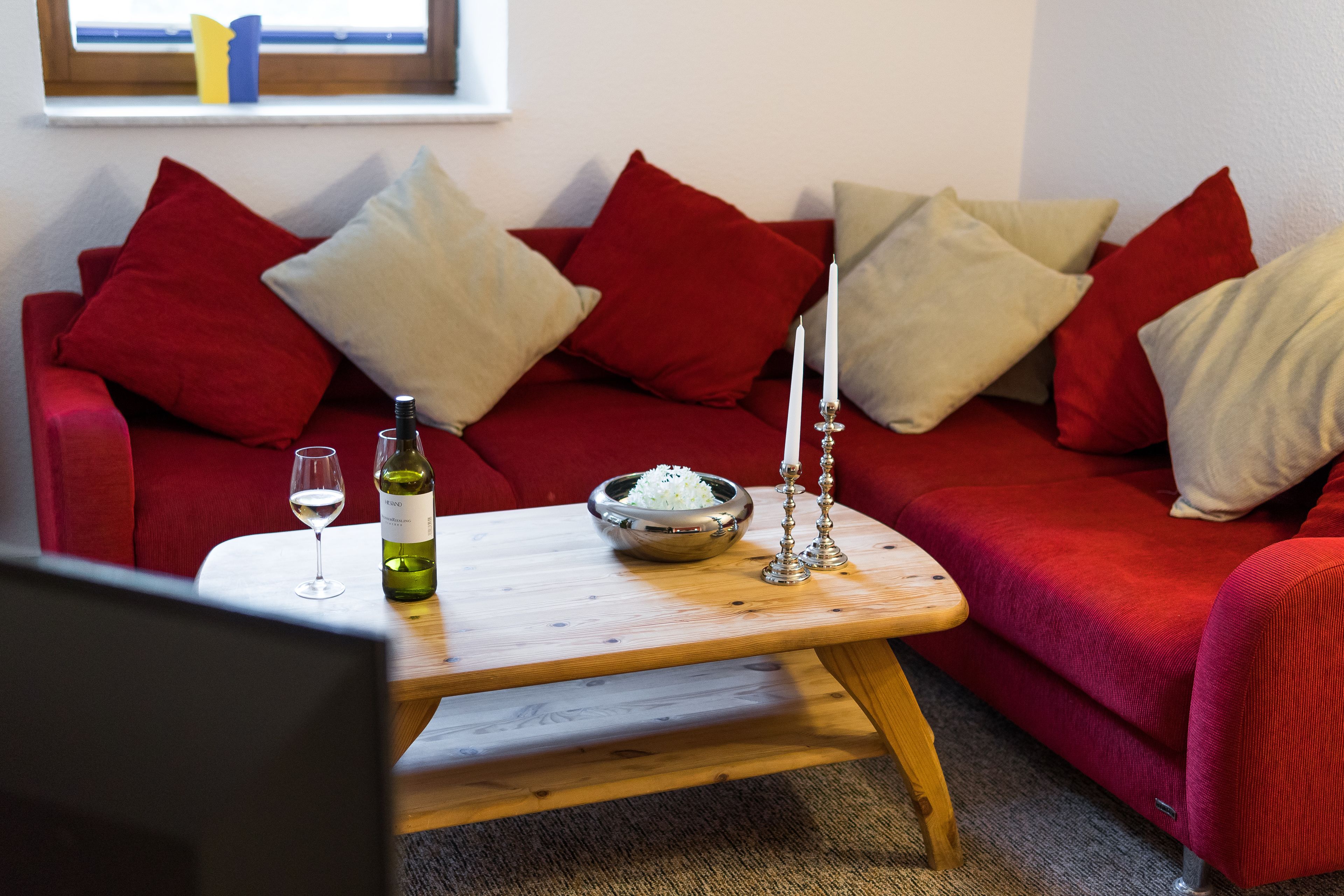 Een rode bank met meerdere kussens, een houten salontafel met een kom rijst, een wijnglas en een fles wijn.