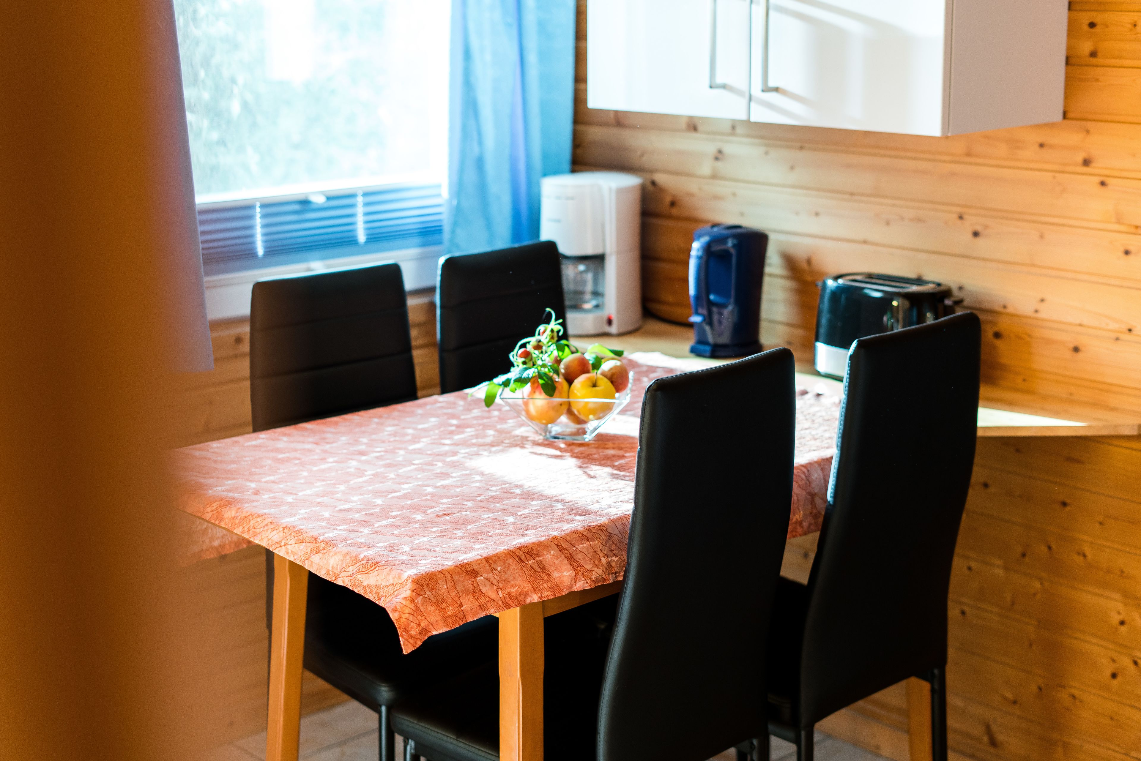 Een eettafel met een rode tafelkleed, zwarte stoelen en een schaal fruit. Op tafel staan een koffiezetapparaat en een broodrooster. De kamer heeft houten wanden en een raam met gordijnen.
