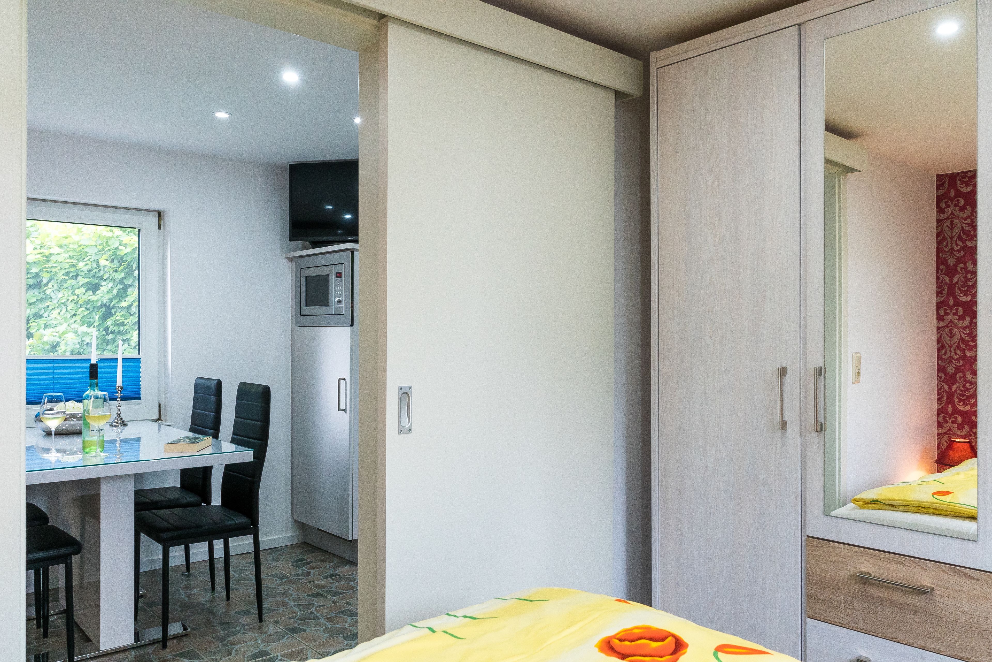 Een kleine kamer met een eethoek, een bed met een geel sprei en een garderobe met spiegel.