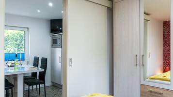 Een kleine kamer met een eethoek, een bed met een geel sprei en een garderobe met spiegel.