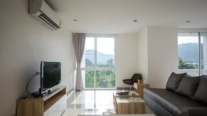 A modern living room with a flat-screen TV, a sofa, a coffee table, and a large window offering a view of greenery and mountains. - Ozone Khaotao (Hua Hin)
