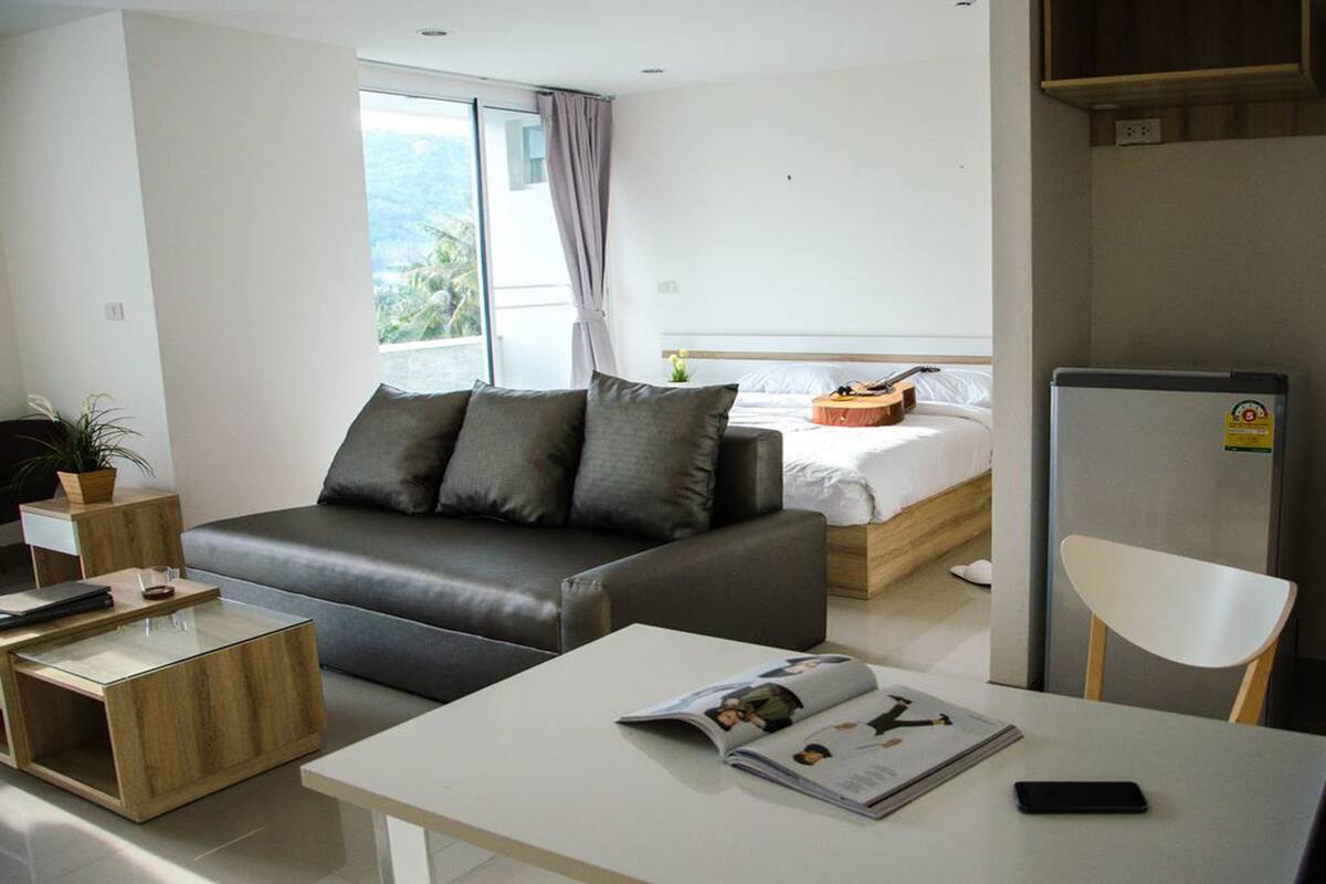 A modern hotel room with a grey sofa, a small wooden side table, a white coffee table, and a bed with a wooden headboard.
