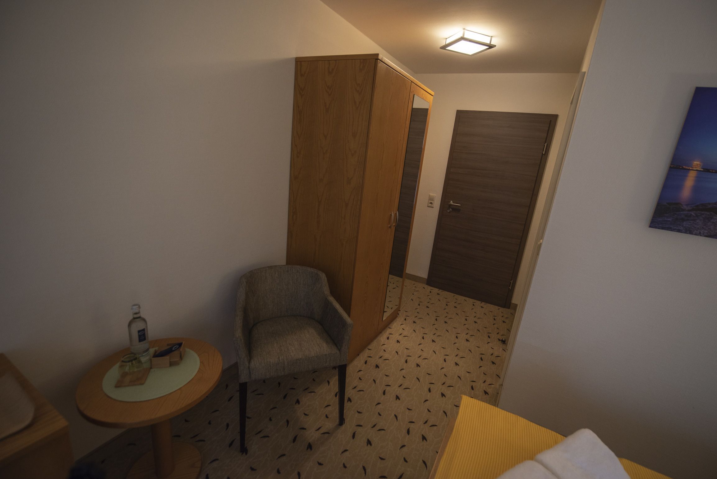A small, dimly lit room with a wooden wardrobe, a chair, a small table with a bottle and glasses, and a door with a view of the outside.