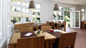 Un vaste espace dédié à la salle à manger, avec des tables et des chaises en bois, de grandes fenêtres équipées de stores et des luminaires suspendus.
