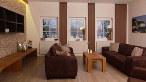 A living room with a brown leather sofa, a wooden coffee table, and a bench. There are three windows with curtains, a brick wall, and a painting on the wall.