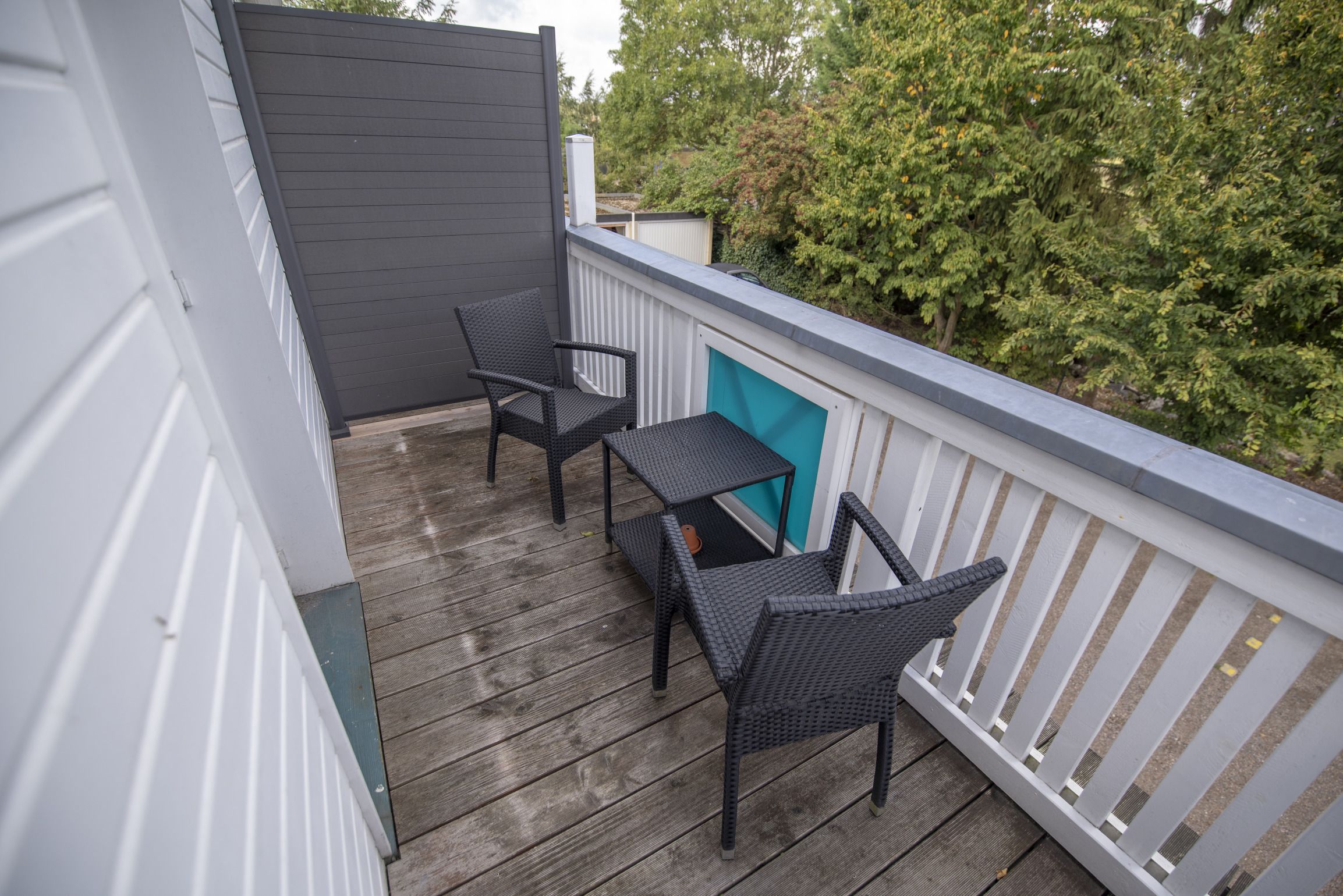 A balcony with two wicker chairs and a small table, wooden deck flooring, and a view of trees and a building in the background.