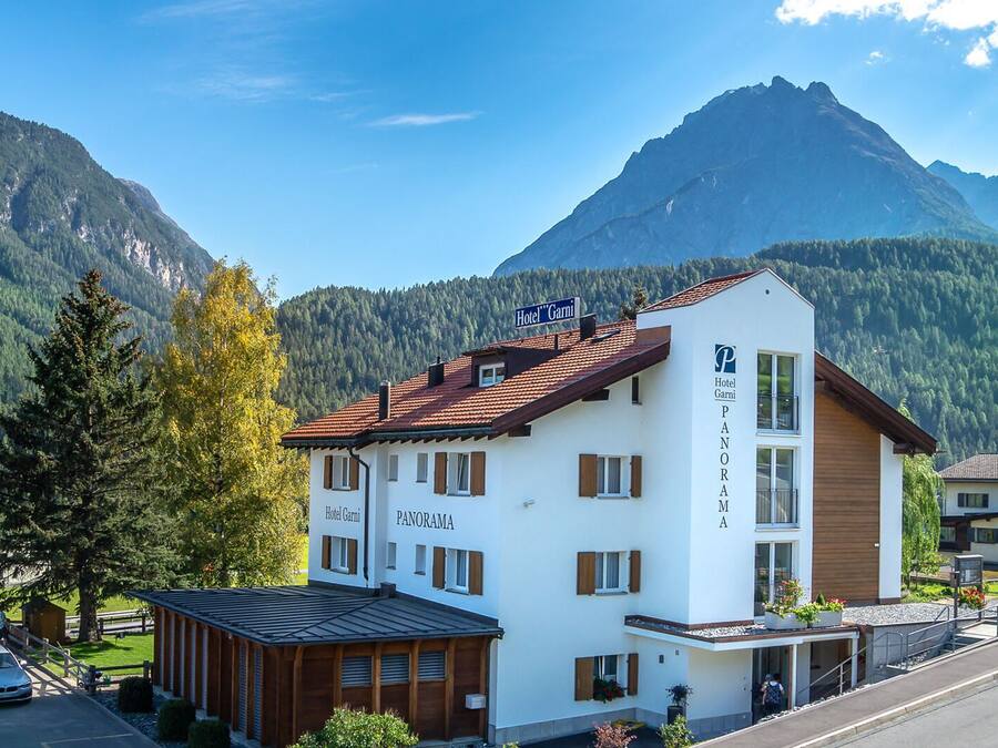 A white building with a red roof, labeled "Hotel Garni Panorama," situated in a mountainous area with a clear sky.