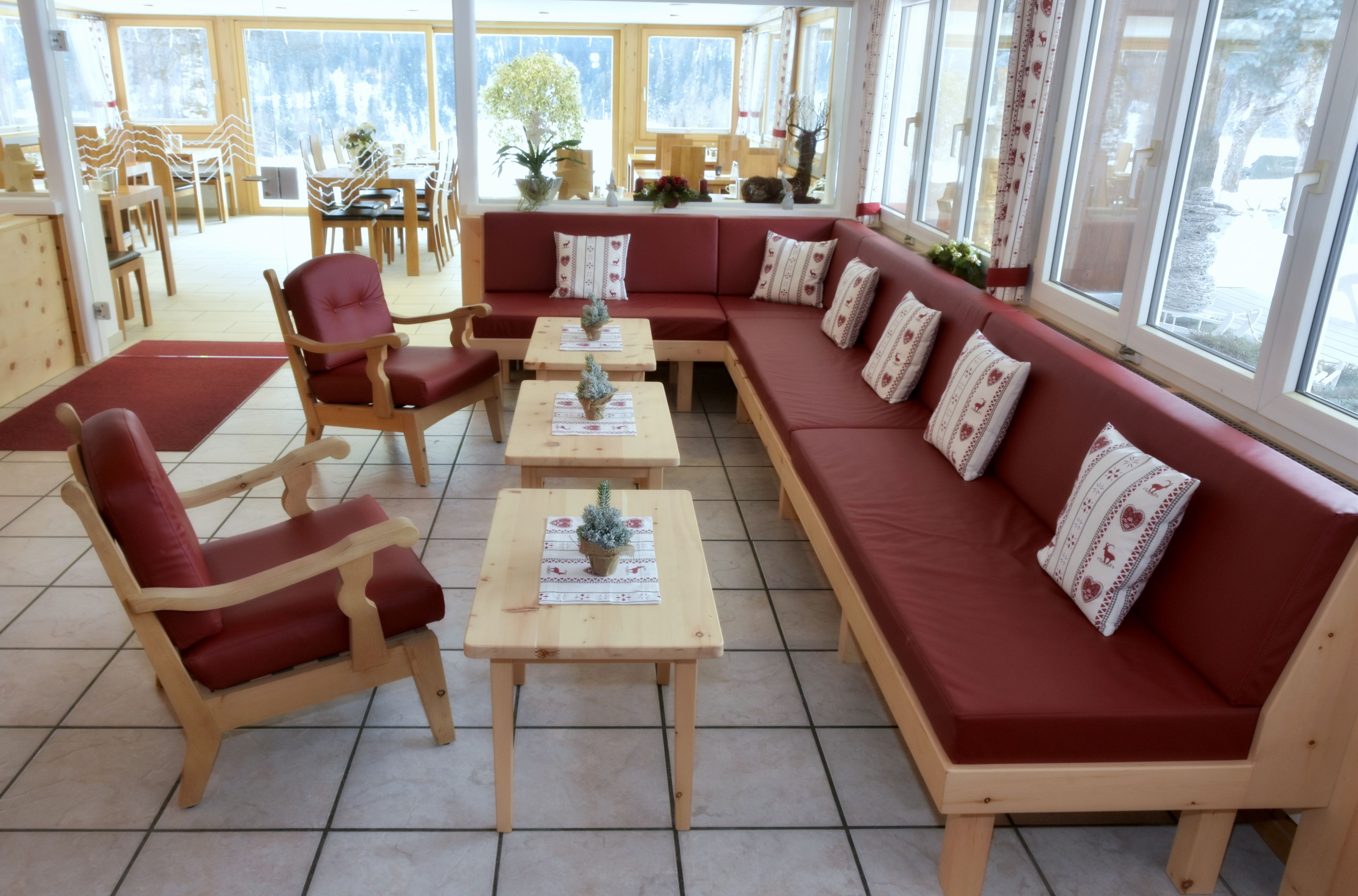 A cozy indoor seating area with a red sofa, wooden chairs, and small tables, featuring large windows and a view of the outdoors.