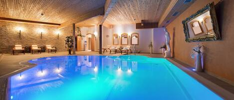 Una piscina interior con techo de madera, sillas de descanso y una estatua.