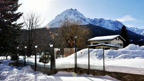 A snow-covered landscape with a mountain in the background, a house with a snow-covered roof, and a street with lampposts and bare trees. - Hotel Panorama (Scuol)