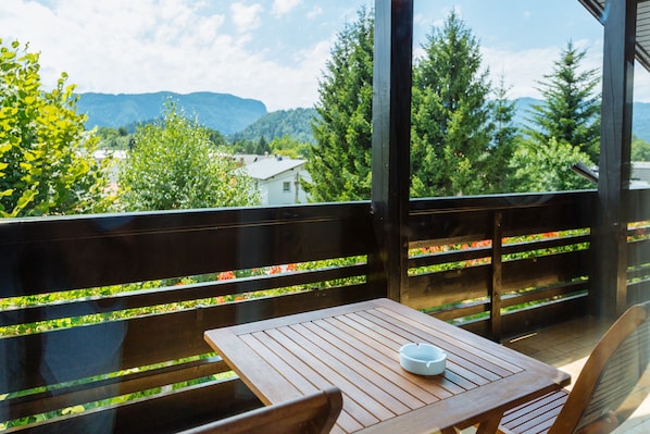 Una terraza de madera con una mesa y sillas, con vistas a un paisaje montañoso, vegetación y una casa al fondo.