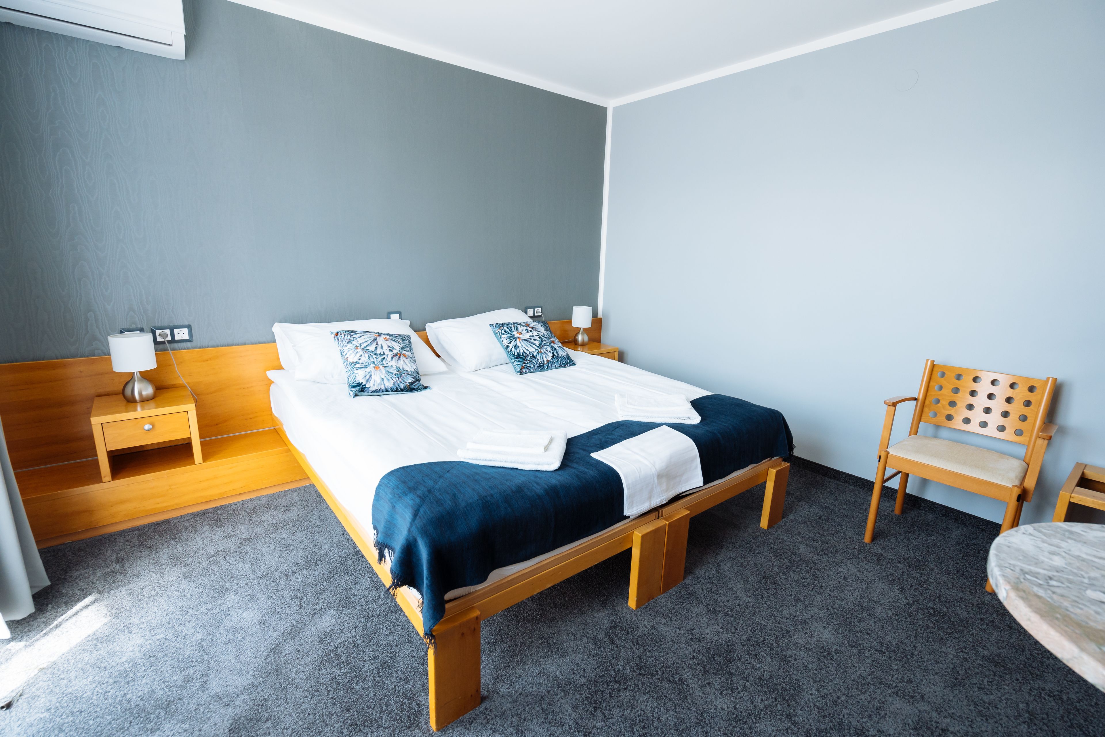 A neatly arranged hotel room with a wooden bed, a wooden chair, a small bedside table, and a nightstand.