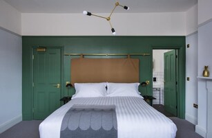 Una cama bien hecha con una colcha a rayas y un cojín decorativo. Arriba, una lámpara de araña moderna, y puertas verdes a cada lado.