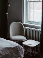 Una habitación con un sillón gris, una pequeña mesa blanca con un documento, un radiador y una ventana con vistas a un edificio de ladrillo.