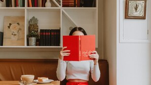 Une personne est assise dans une pièce, lisant un livre rouge. On y voit une bibliothèque, une table en bois avec une tasse et une assiette, et une horloge murale.