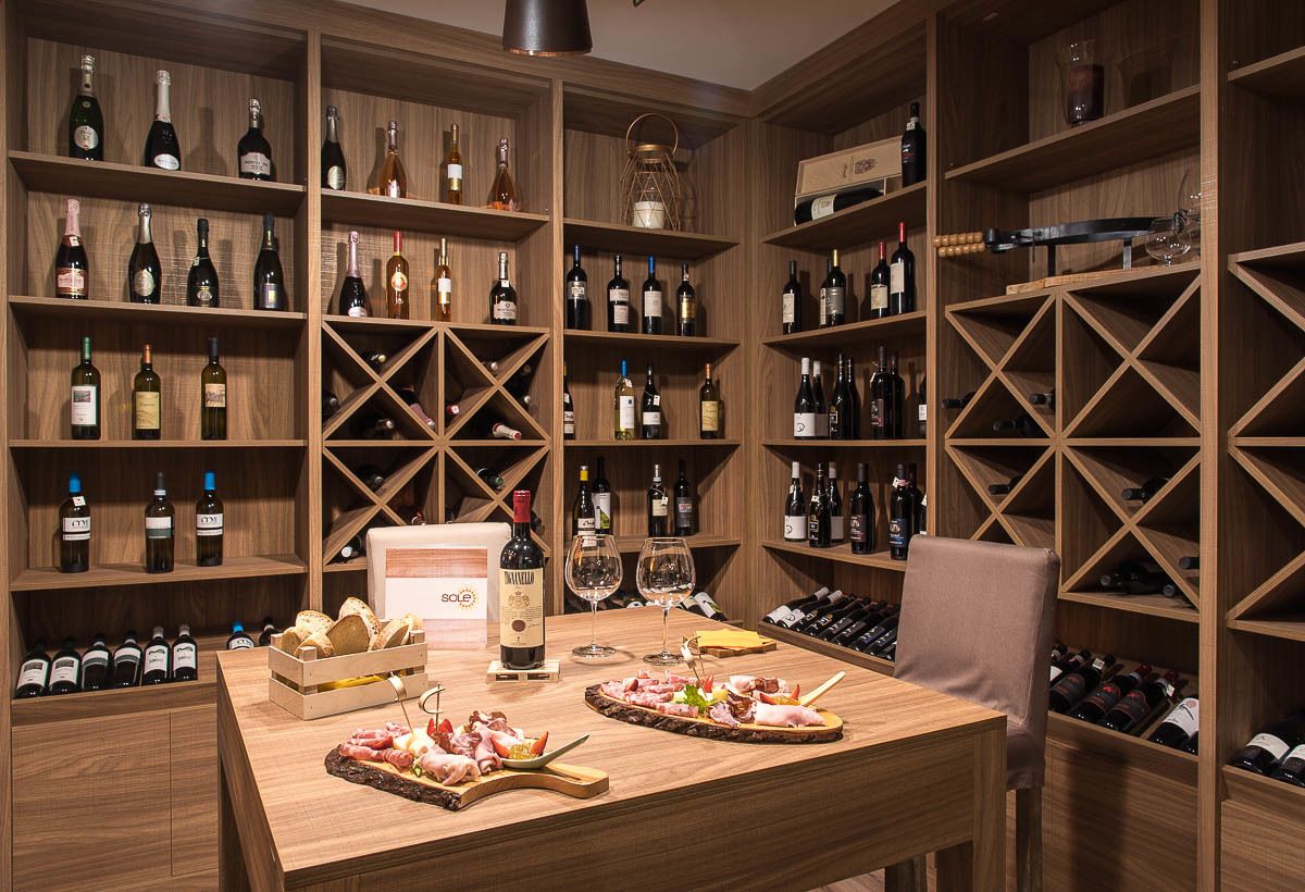 A wine cellar with wooden shelves stocked with various bottles, a dining table with a charcuterie board and glasses, and a chair.