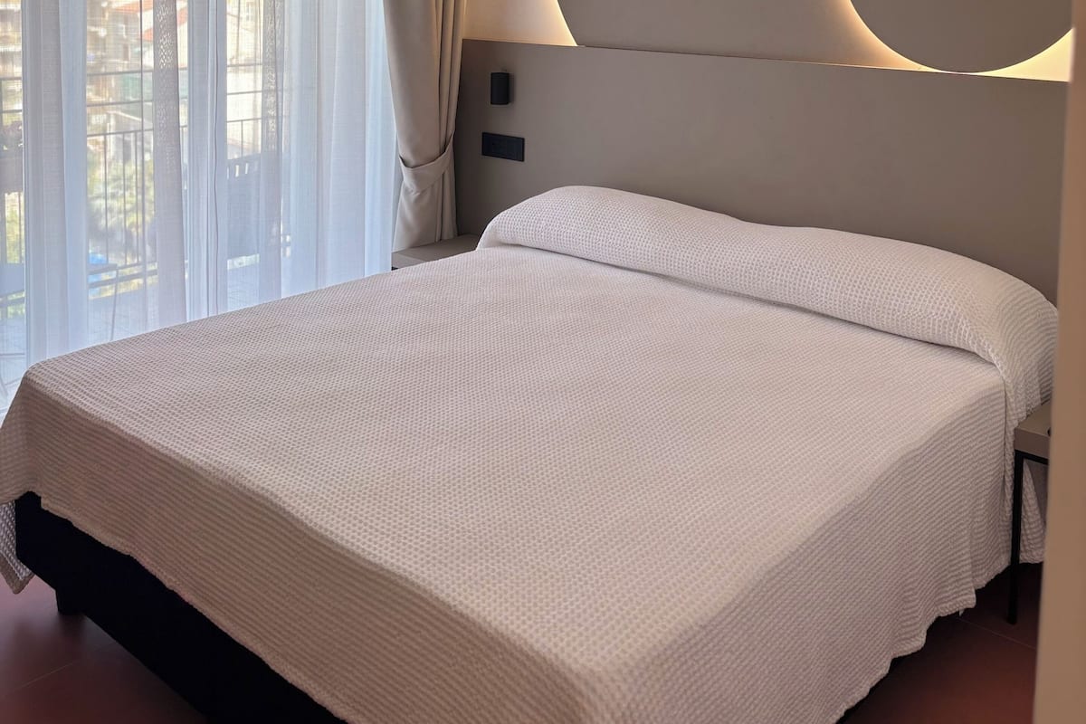 Une chambre d’hôtel avec un grand lit, un applique mural circulaire et des rideaux en voile.