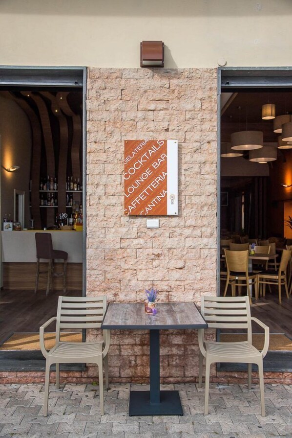 An outdoor seating area with two tables and chairs, a stone wall with a menu sign, and a bar visible through an open door. - Hotel Sole (Maiori)