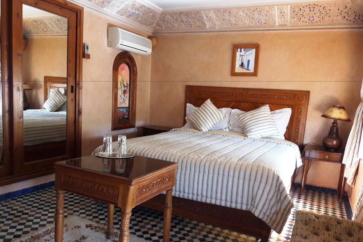 A hotel room with a checkered floor, a wooden bed with striped bedding, a wooden nightstand with a lamp, and a mirror on the wall.