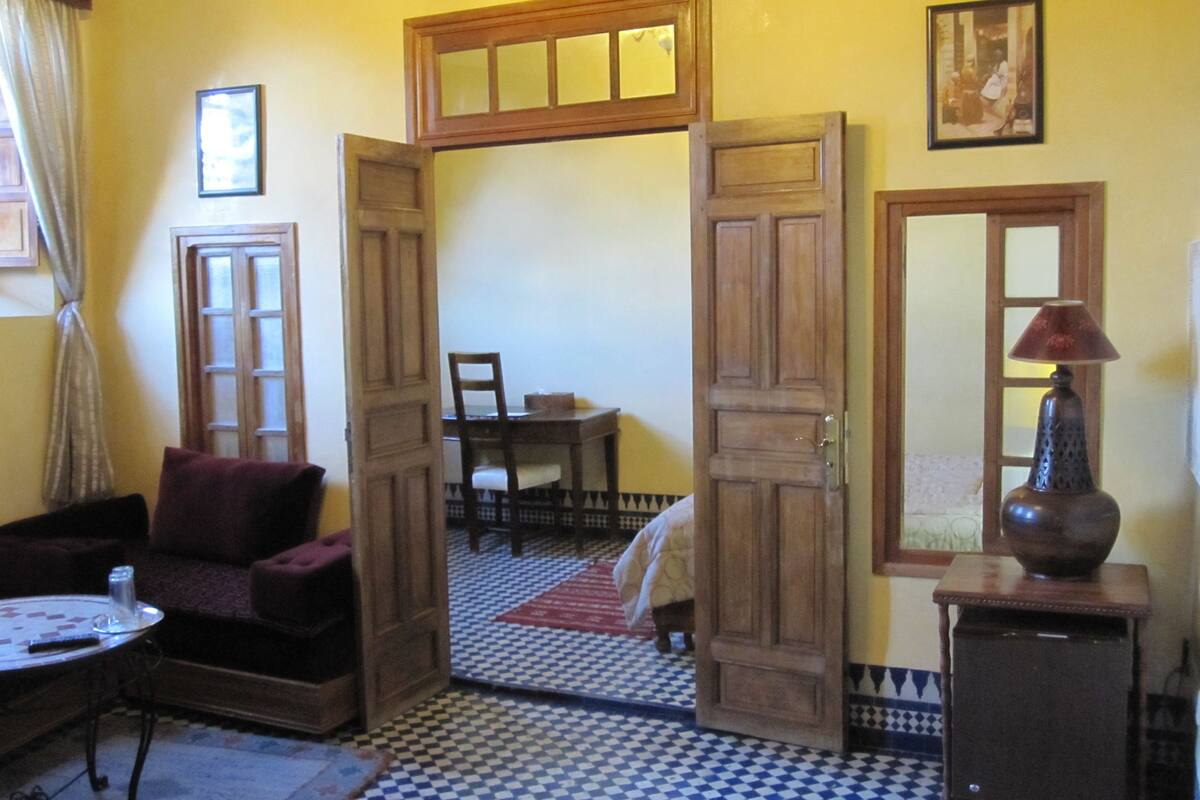 A room with a checkered floor, wooden doors, a dining table with chairs, a sofa, a lamp, and a small cabinet.