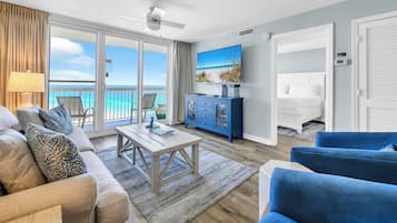 A spacious living room with a large sliding glass door leading to a balcony with a view of the ocean. There is a blue cabinet, a white sofa, and a blue armchair.