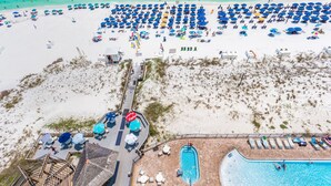 An aerial view of a beach with numerous umbrellas and lounge chairs, a swimming pool, and a wooden deck area. - Pelican Beach Resort by Colasan (Destin)
