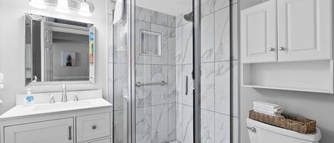 A modern bathroom with a glass-enclosed shower, white marble tiles, a large mirror, and white cabinetry.