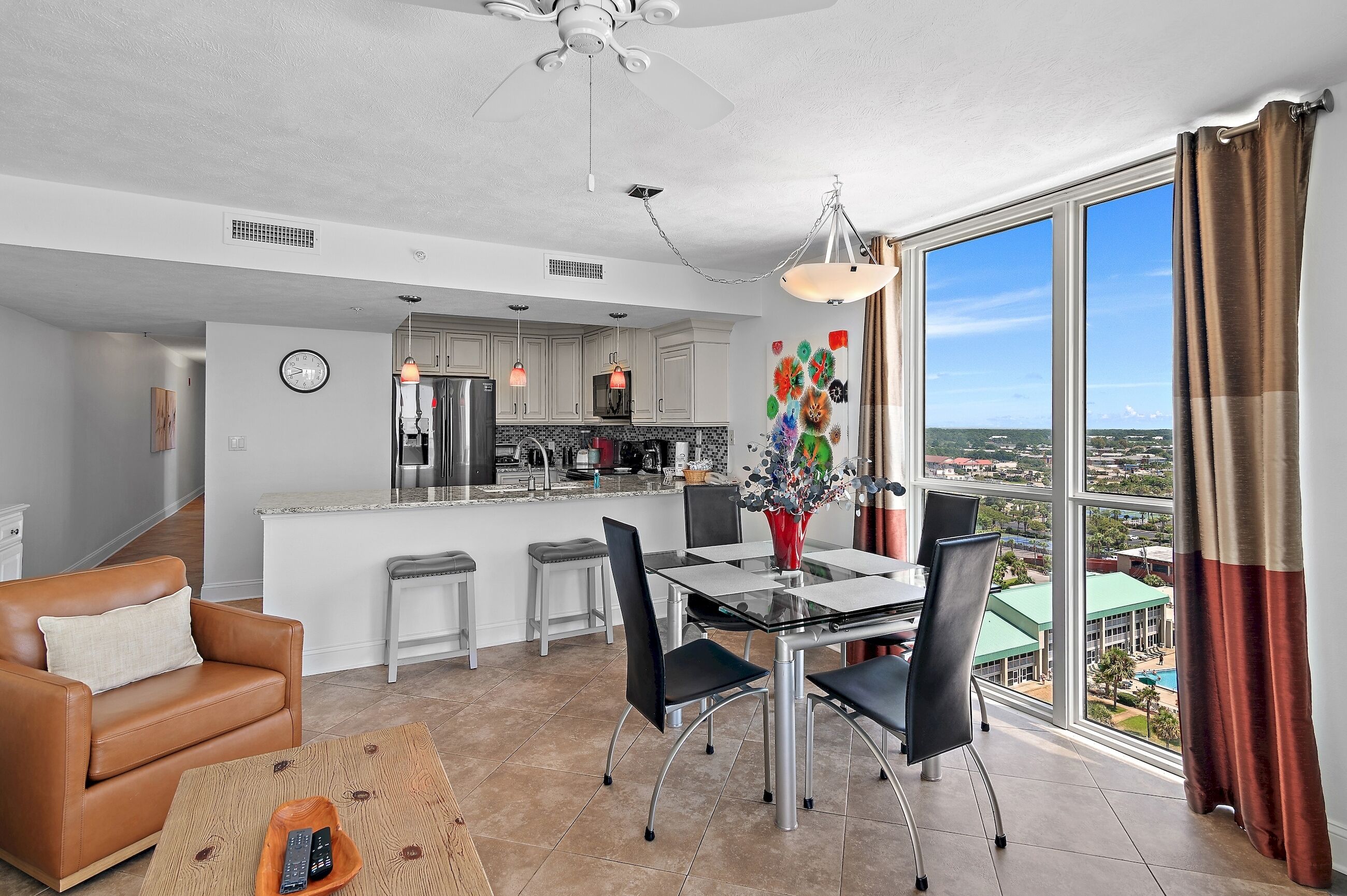 A modern kitchen with white cabinets, a dining area with a glass table and black chairs, a brown leather sofa, and a view of the city through large windows.