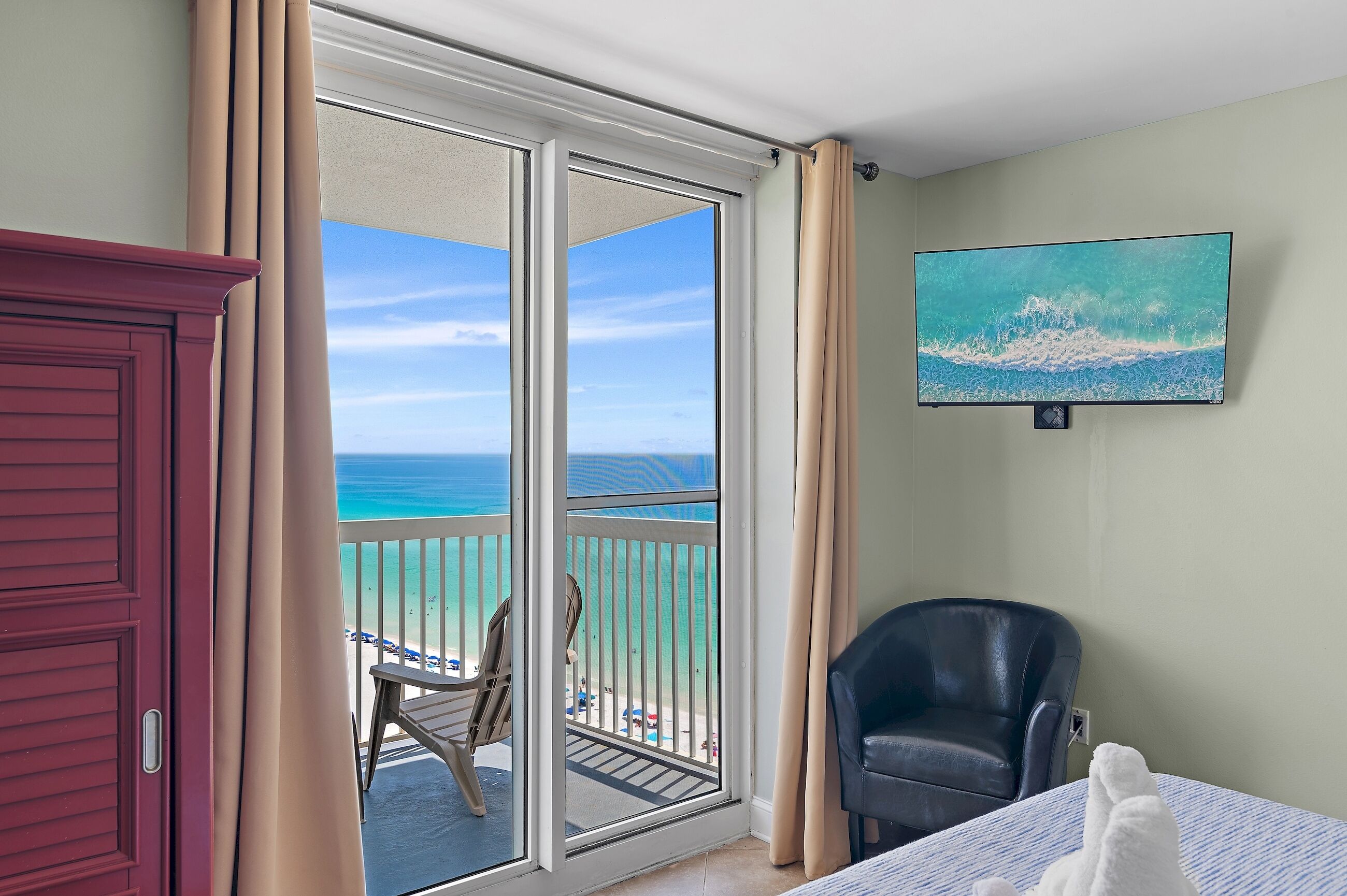 A room with a large sliding glass door leading to a balcony with a view of the ocean. There is a bed with a blue and white striped bedspread, a black leather chair, and a painting on the wall.