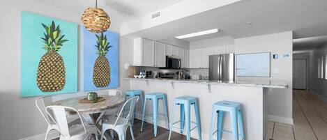 Una cocina moderna con armarios blancos, un refrigerador de acero inoxidable y un microondas. Hay un comedor con una mesa redonda y taburetes de bar azules.