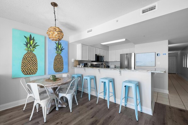 Una cocina moderna con armarios blancos, un refrigerador de acero inoxidable y un microondas. Hay un comedor con una mesa redonda y taburetes de bar azules.