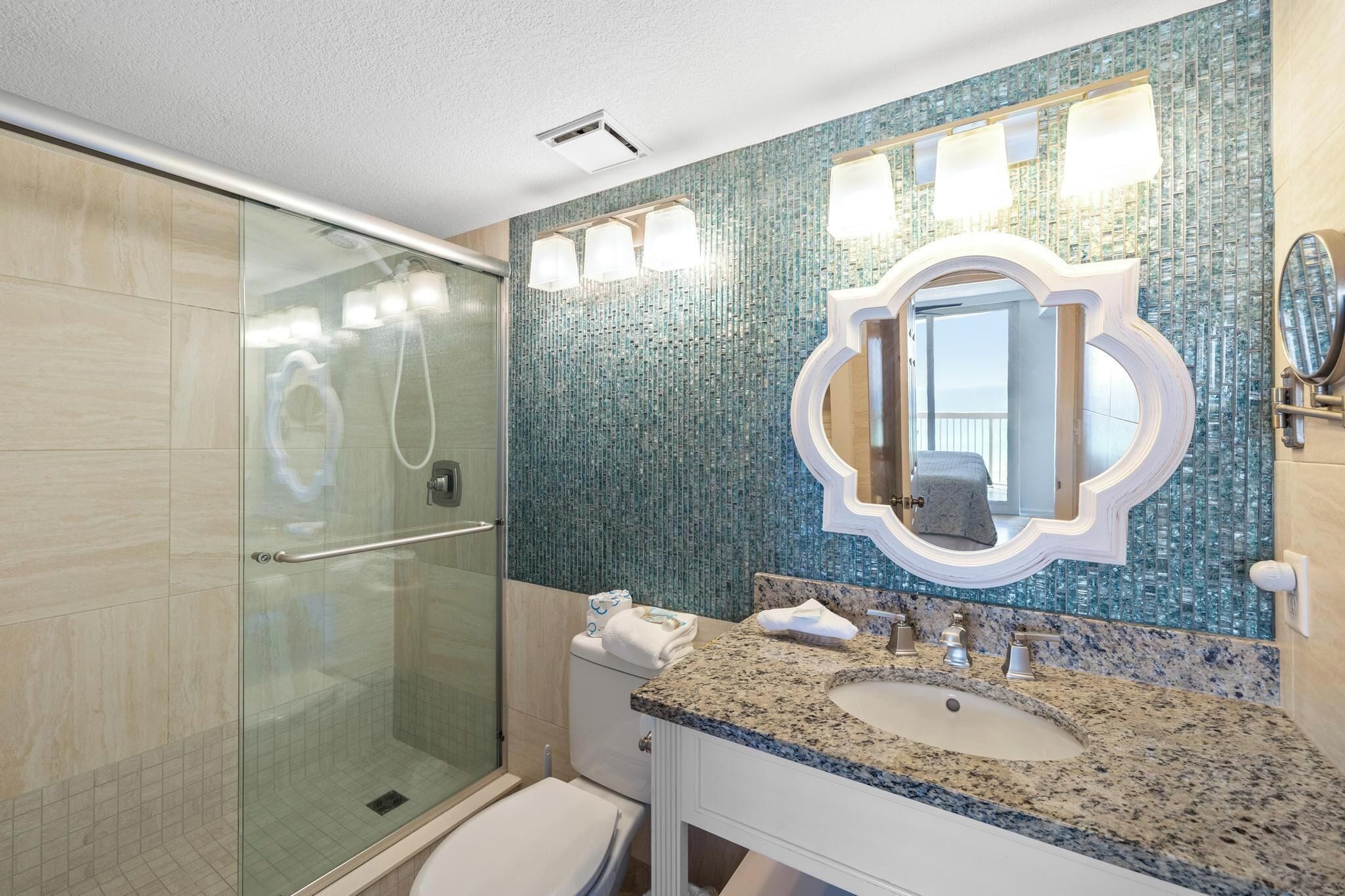 A bathroom with a glass shower enclosure, a sink with a granite countertop, and a mirror with a decorative frame.