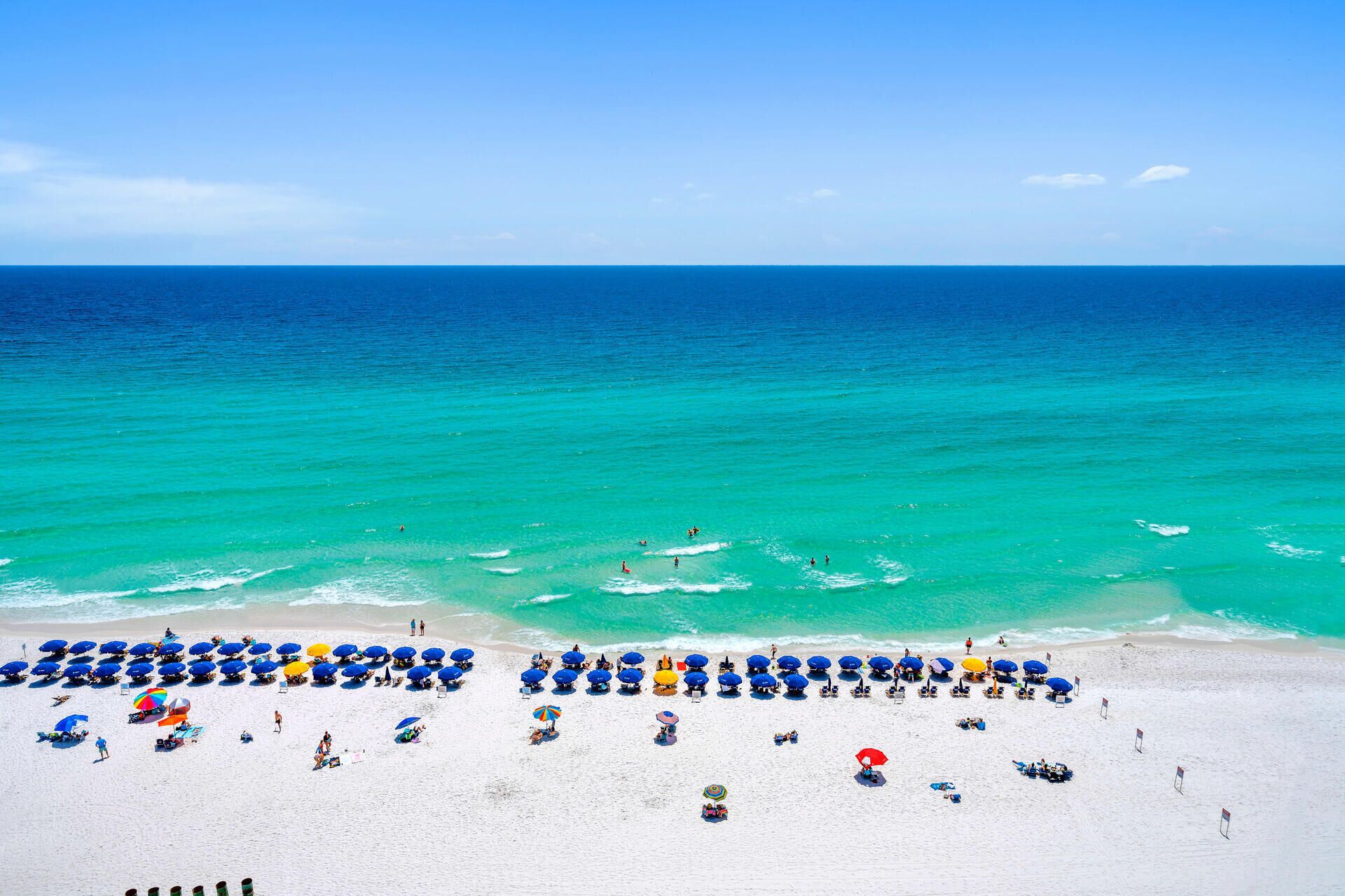 A beach with rows of umbrellas and beach chairs, a clear blue ocean, and a sandy shore.