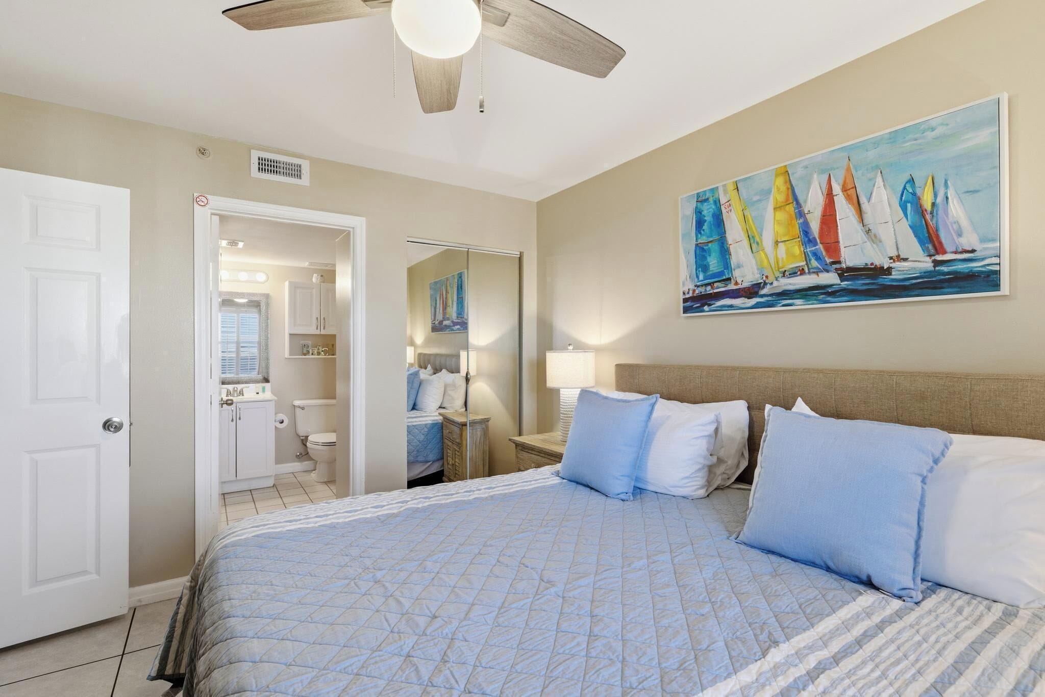 A bedroom with a bed, a ceiling fan, a painting of sailboats, and a bathroom visible through an open door.