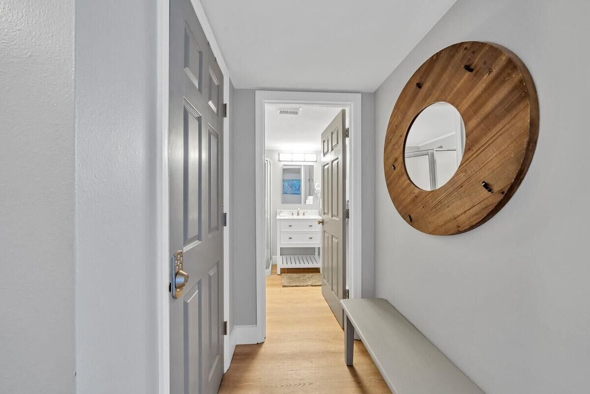 A hallway with a wooden mirror, a bench, and a view into a bathroom with a sink and mirror.
