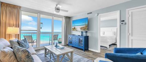 Een ruime woonkamer met een groot raam met uitzicht op een strand, een plafondventilator, een televisie en een bed in de aangrenzende kamer.