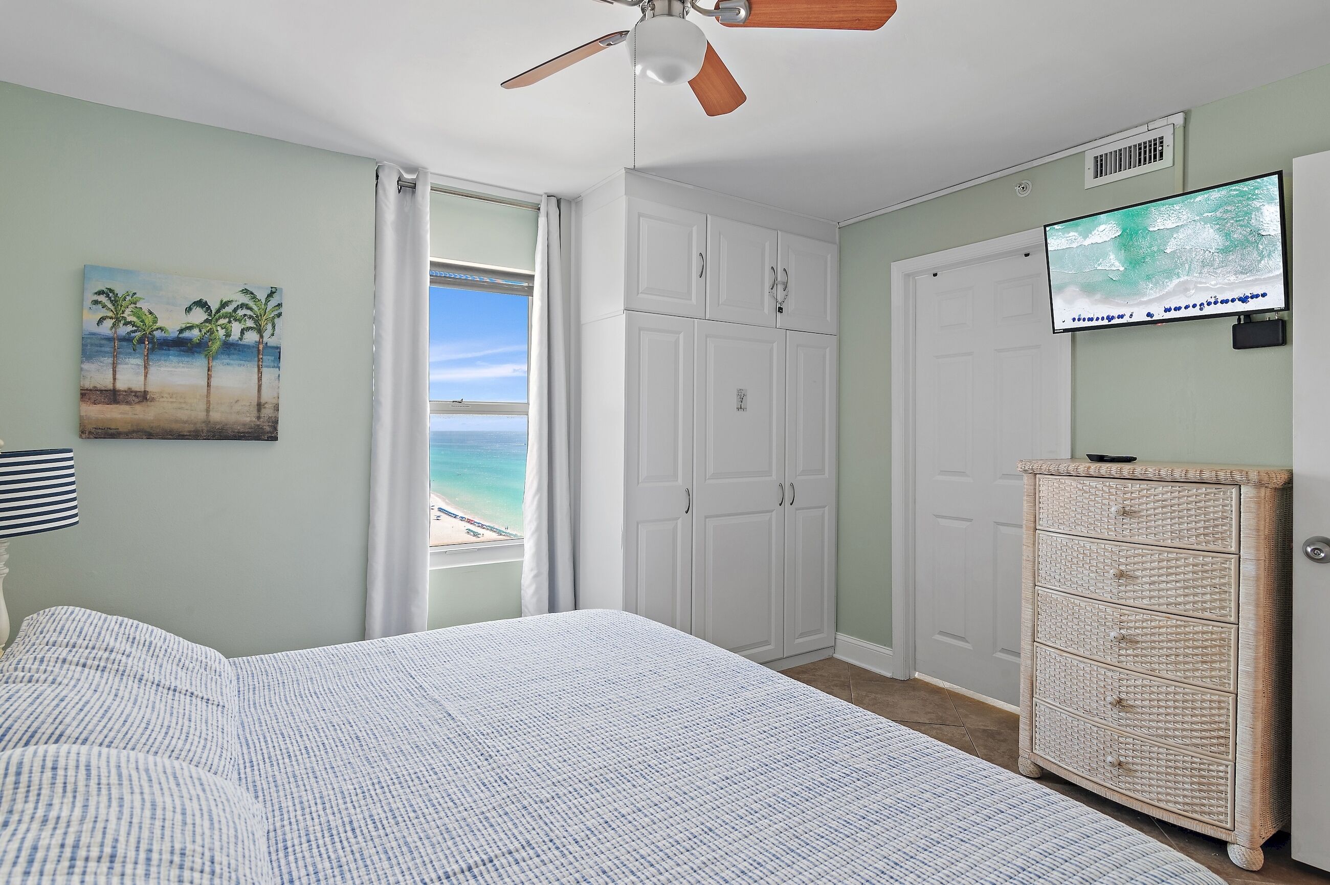A bedroom with a bed, a ceiling fan, a TV, a wardrobe, and a painting of a beach scene.