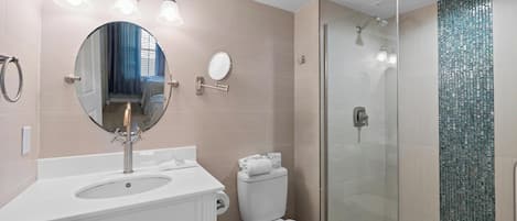 Un baño moderno con lavamanos, espejo, inodoro y ducha con mampara de vidrio.