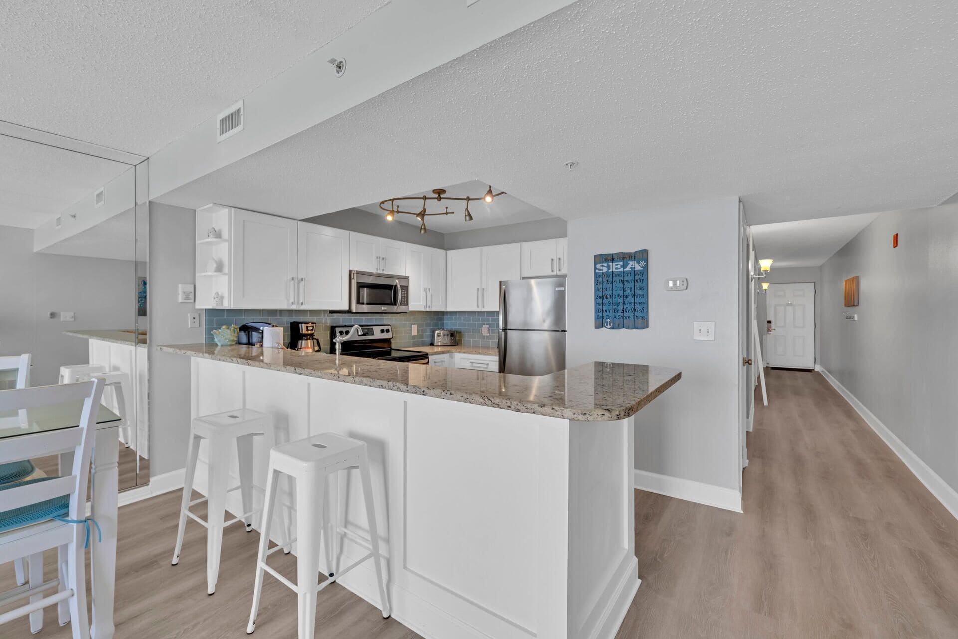 A modern kitchen with white cabinetry, a granite countertop, and stainless steel appliances. There are two white bar stools and a dining table with chairs.