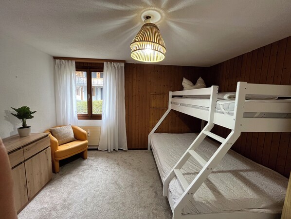 A bunk bed room with a wooden ceiling, a window with curtains, a small table with a plant, and a hanging light fixture.
