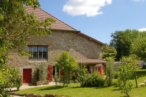 Un bâtiment en pierre avec un toit recouvert de tuiles rouges, un petit porche et un jardin bien entretenu.