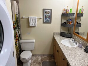 Ein Badezimmer mit Toilette, Waschbecken und einem Handtuchhalter mit Handtuch. An der Wand hängt ein gerahmtes Bild und eine Ablage mit Pflegeprodukten.