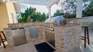 An outdoor kitchen with a built-in grill, a refrigerator, and a sink, surrounded by stone walls and a pergola. - Savhaven - 4 Bed, 4 Bath with Pool, Sleeps 8 (2 minutes away from Spotts Beach) (Savannah)