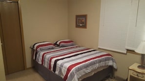 A bedroom with a bed, a nightstand, a window with blinds, and a door. - Lake Cabin for Rent on the Shores of Beautiful Devils Lake, ND. (Devils Lake)