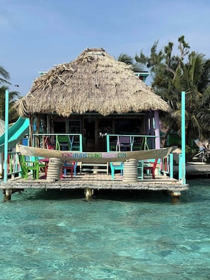 Una estructura con techo de paja, un letrero, una plataforma flotante con sillas y agua turquesa cristalina.