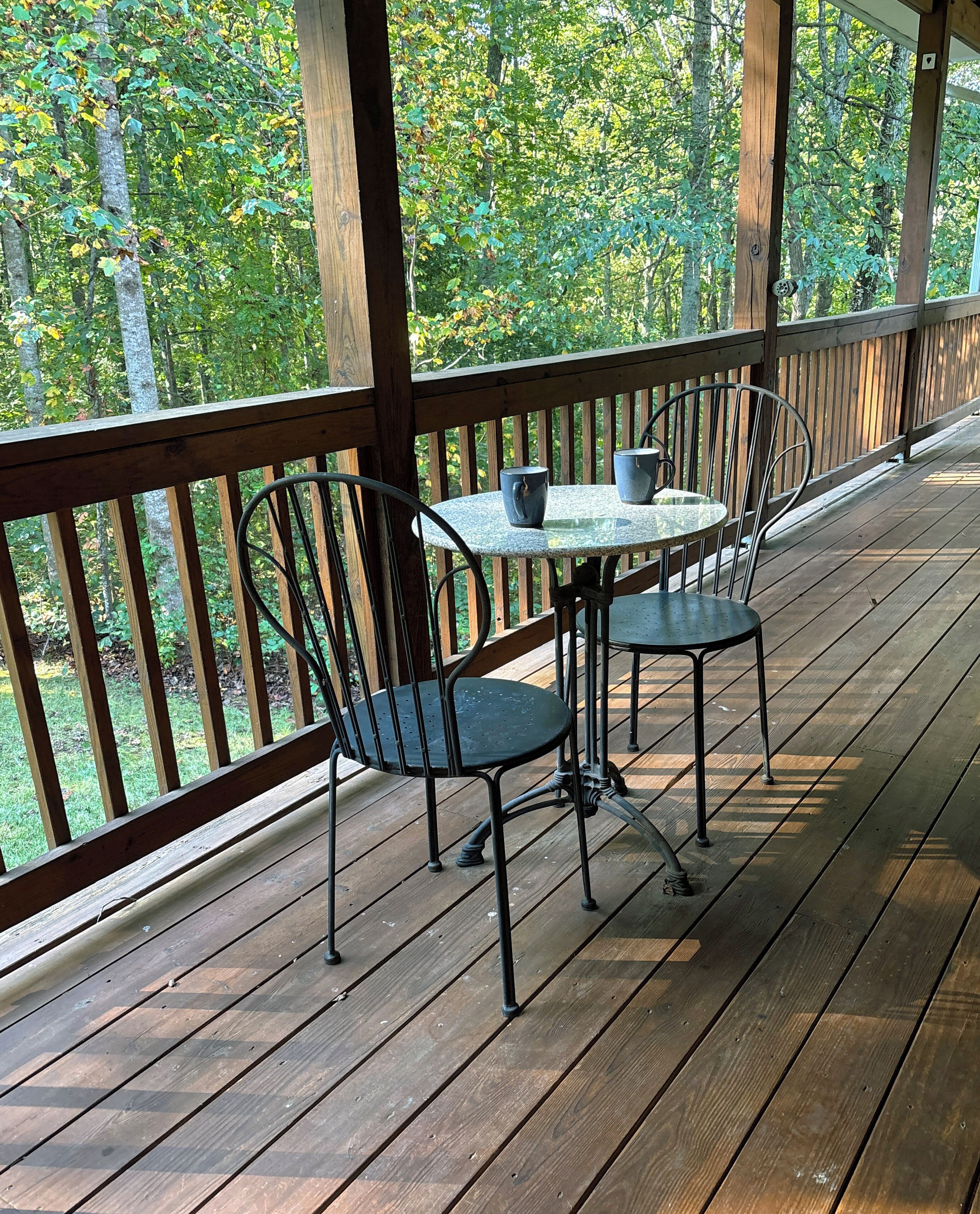 A wooden deck with a table and chairs, surrounded by a railing, overlooking a forested area.