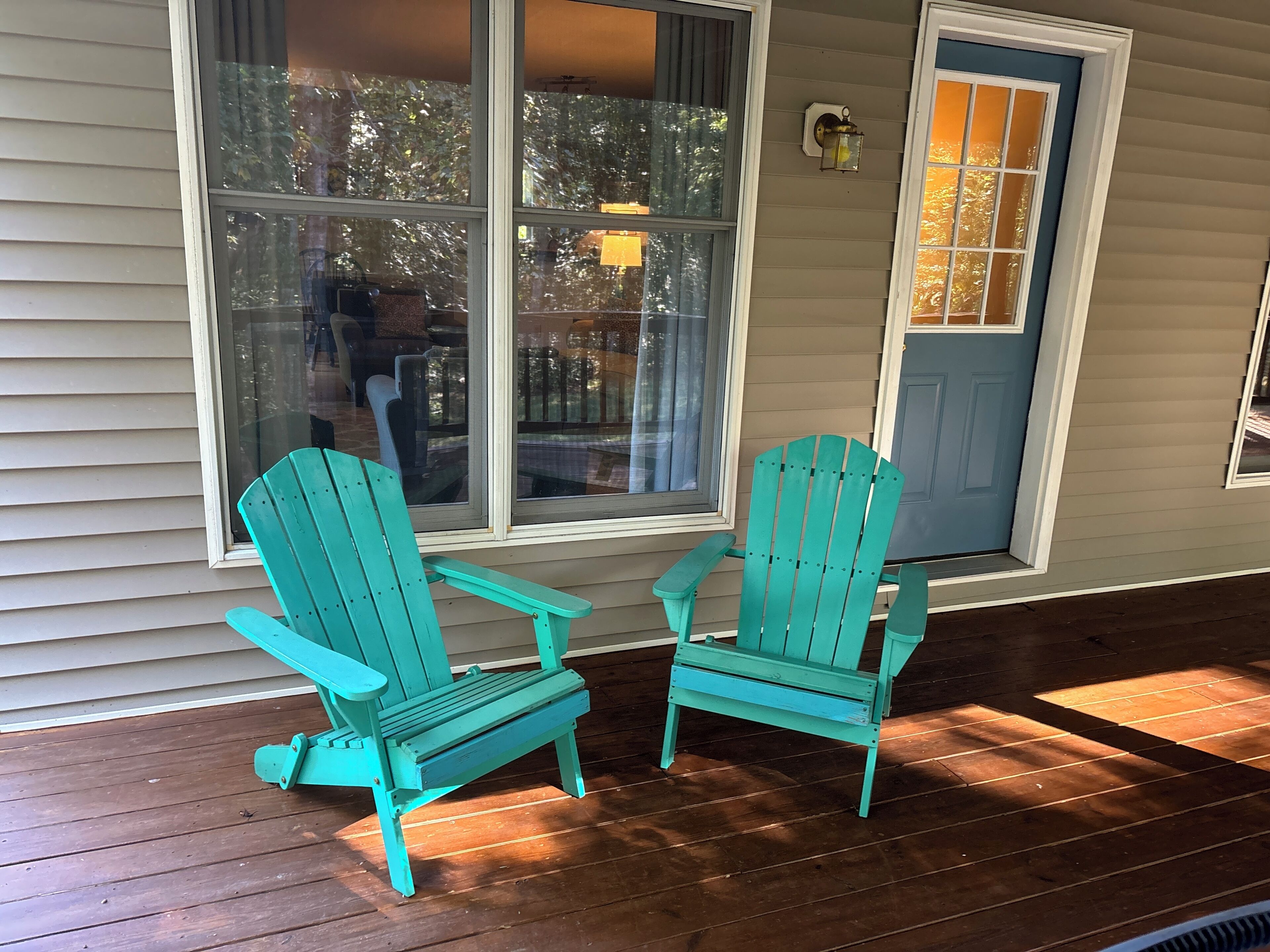 Dos sillas Adirondack turquesa en una terraza de madera frente a una casa con revestimiento beige y una puerta azul.