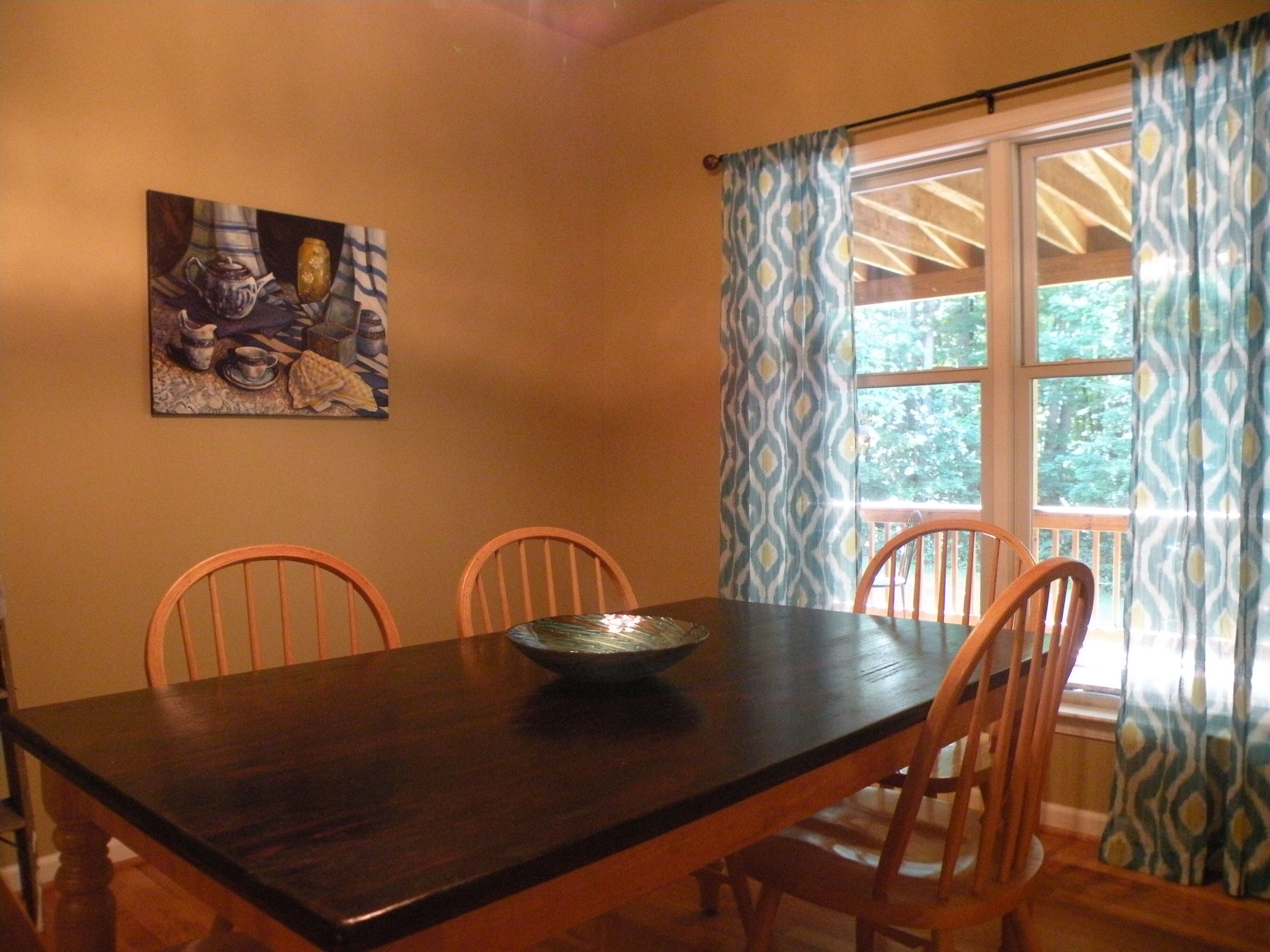 Un comedor con una mesa de madera oscura, cuatro sillas de madera, un bol en la mesa y una ventana con cortinas estampadas.