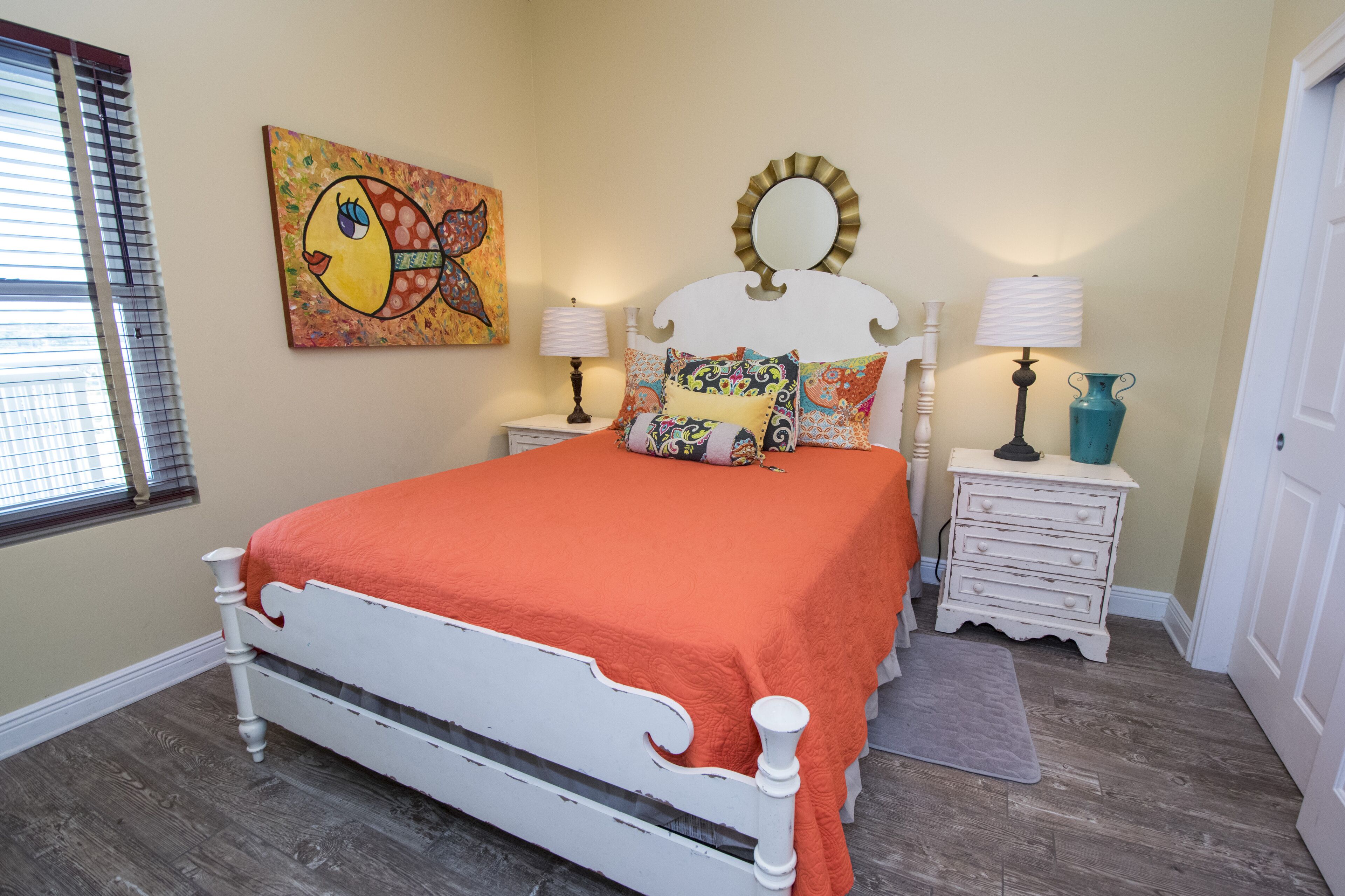 A bedroom with a white bed, orange bedding, a colorful painting, two bedside tables with lamps, and a wooden floor.