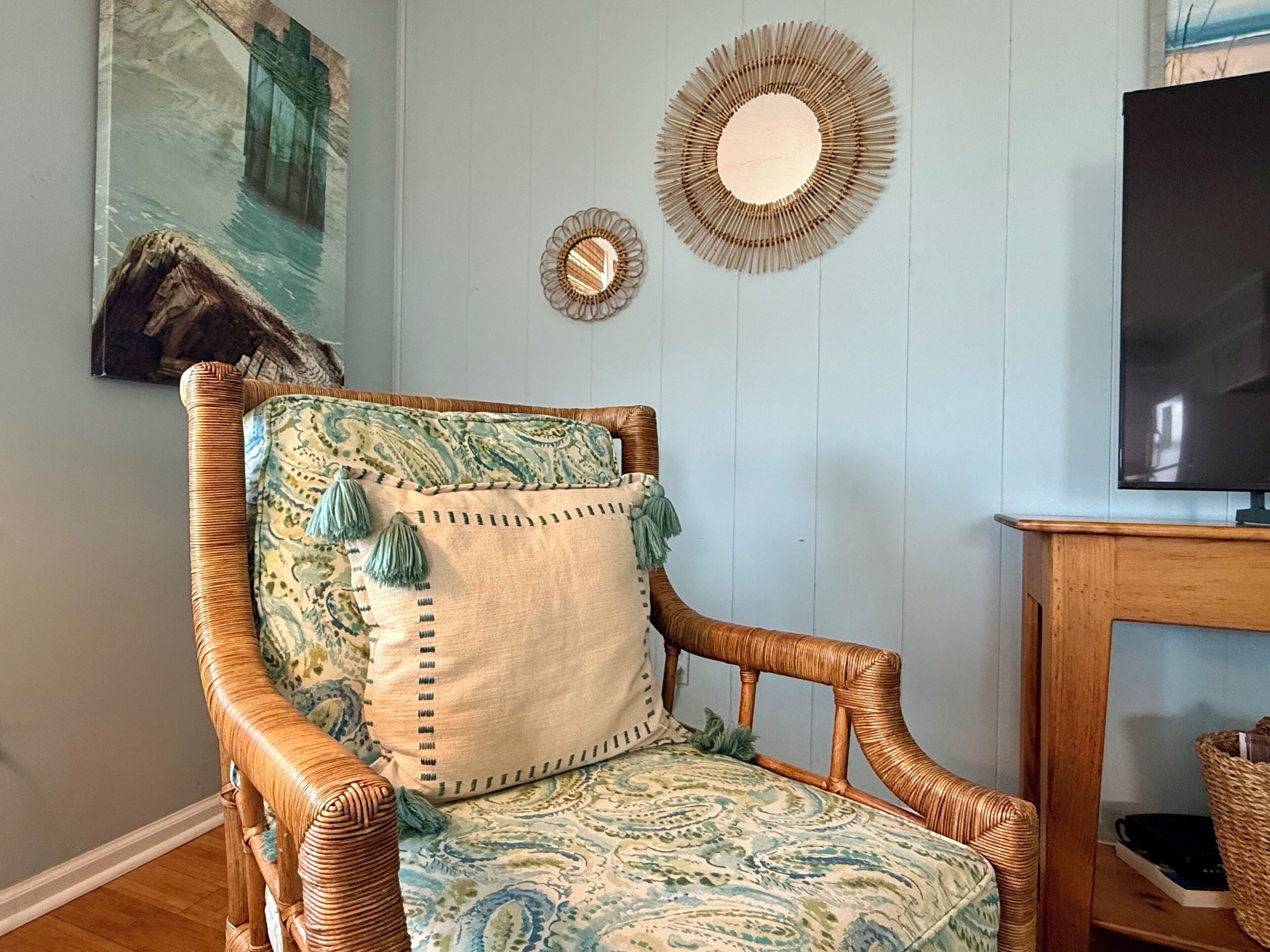 Une chaise en osier avec un coussin à motifs et des glands décoratifs, un miroir rond et un téléviseur posé sur un meuble en bois.